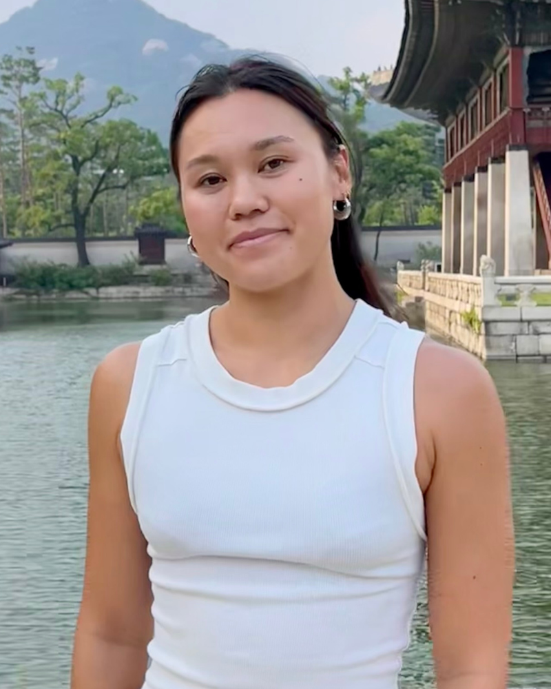 Portrait of a woman wearing a white sleeveless top standing outdoors beside a body of water, with a traditional building and mountains in the background.